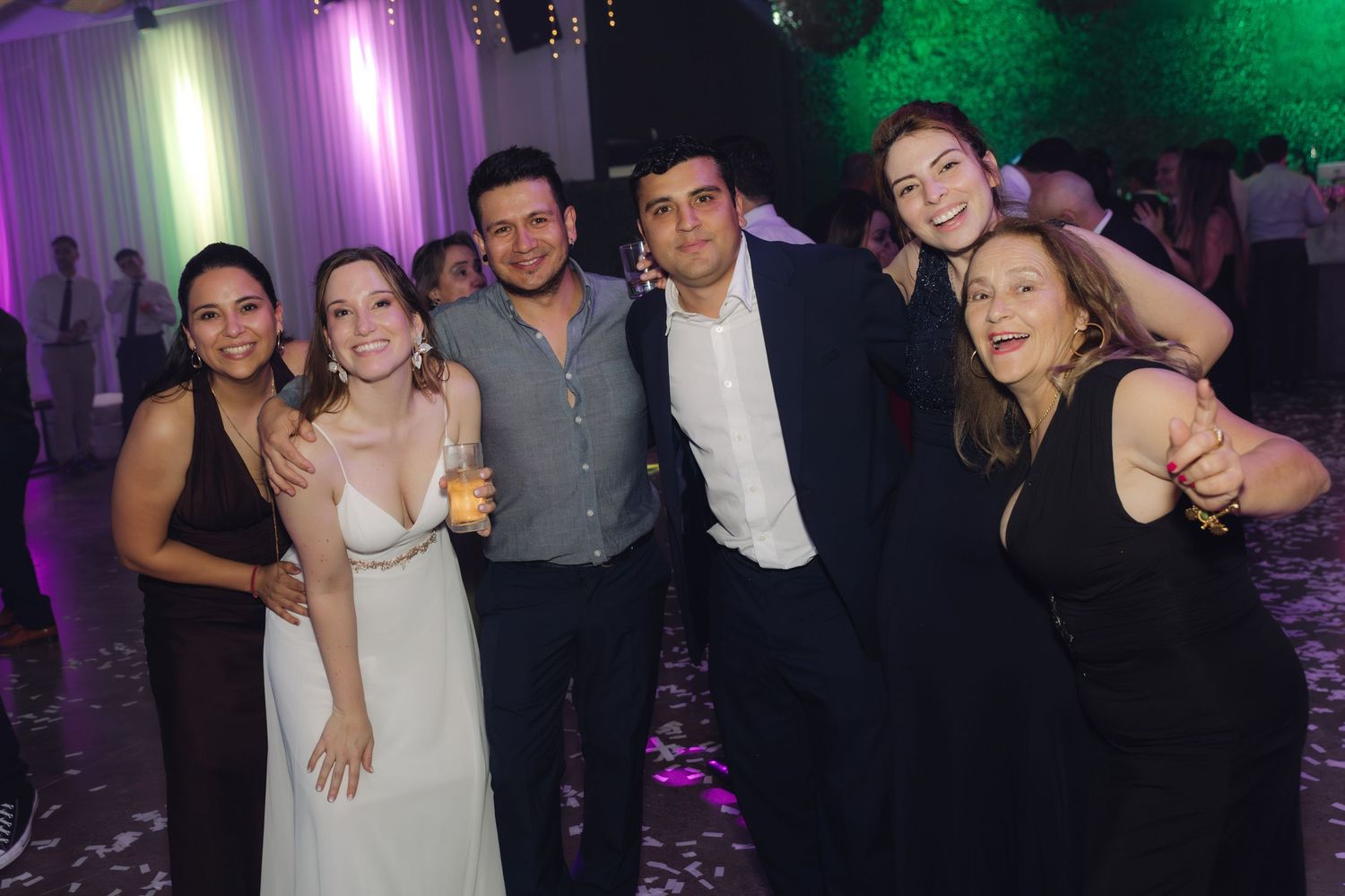 Bride in white dress poses with five smiling friends at vibrant wedding reception party.