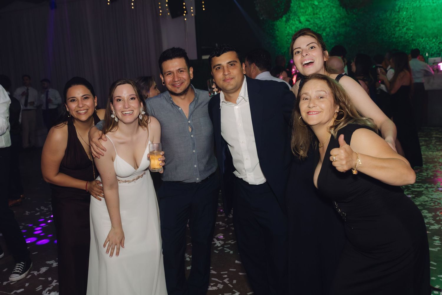 Bride and five guests smile together on colorful lit dance floor at wedding celebration.