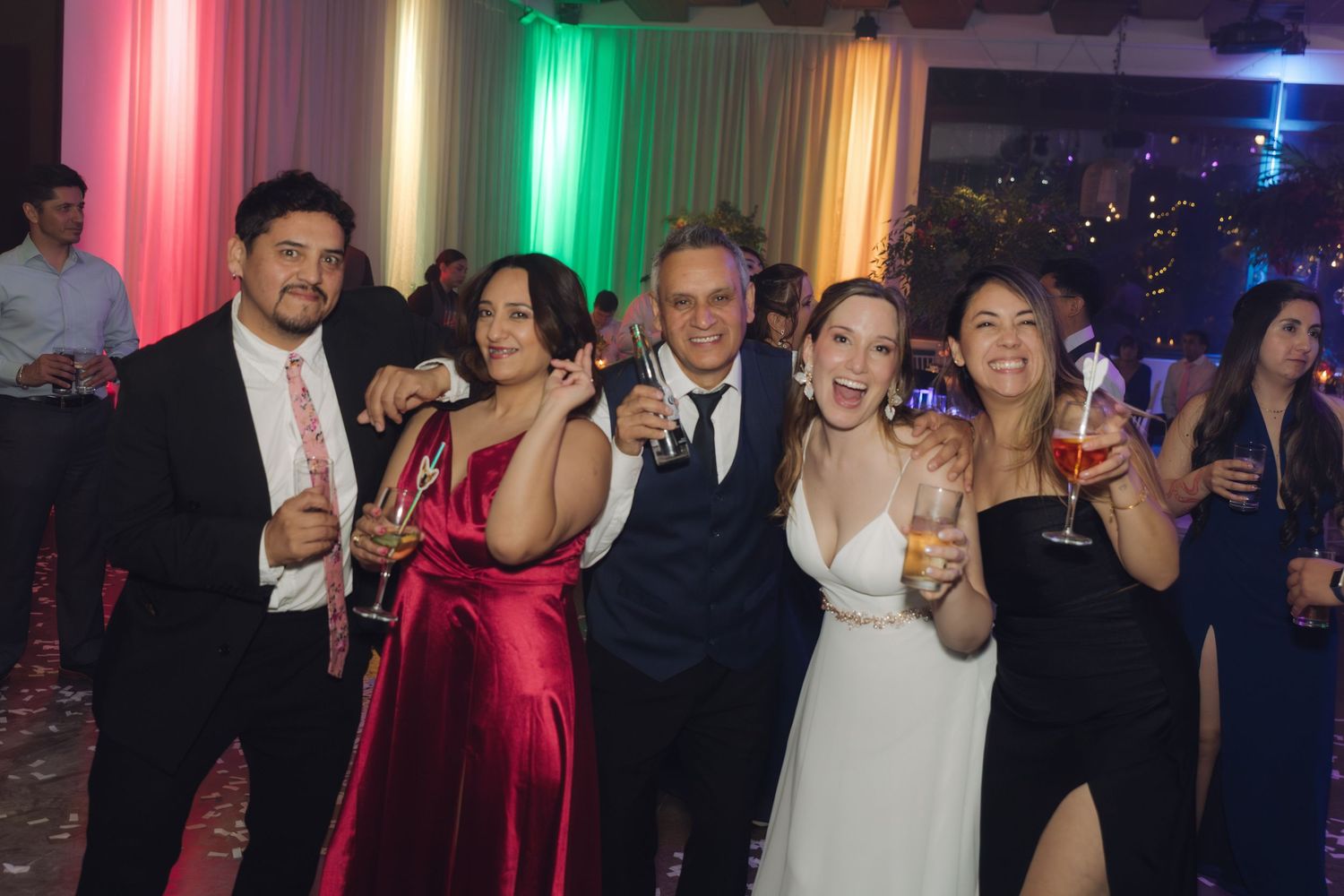 Bride in white gown poses with four guests holding drinks at festive wedding reception.