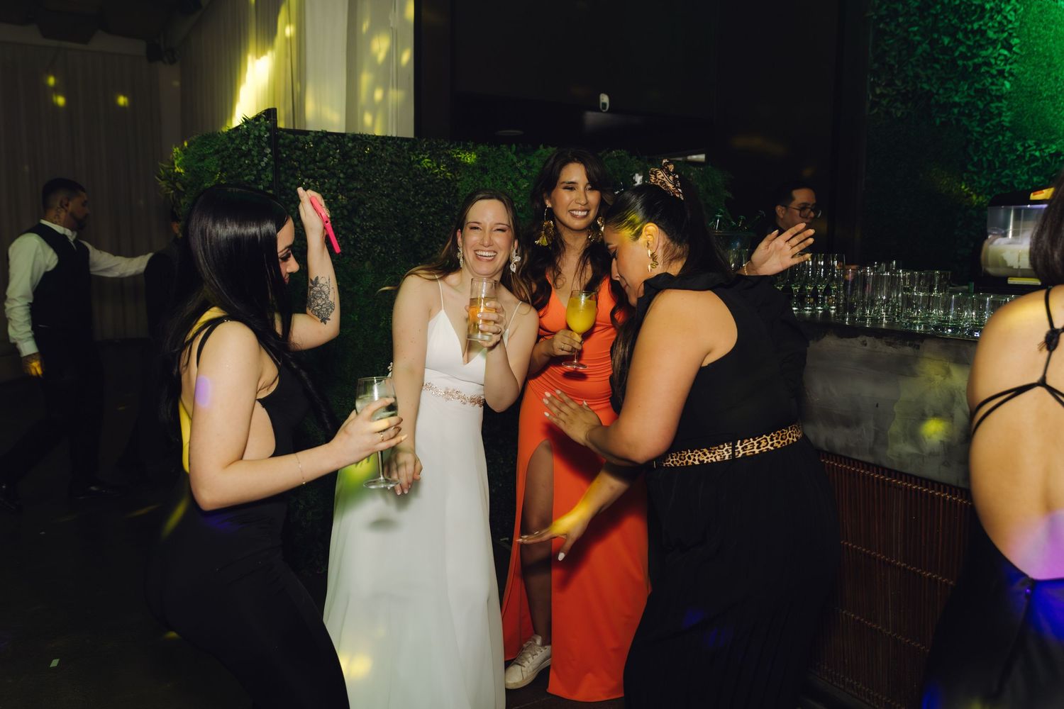 Bride in white dress laughs with friends near bar at elegant evening wedding reception.