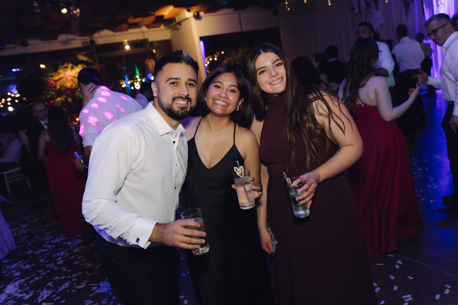 Three guests smile and pose with drinks at elegant evening wedding reception celebration.
