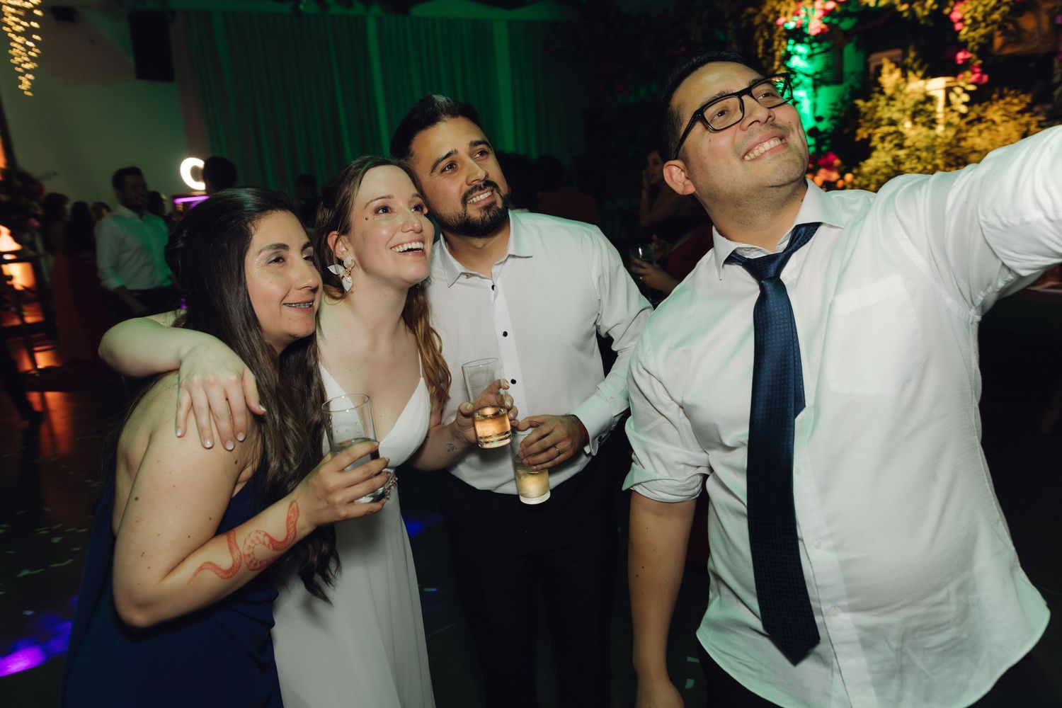 Four wedding guests smile and take a selfie while holding drinks on the dance floor.