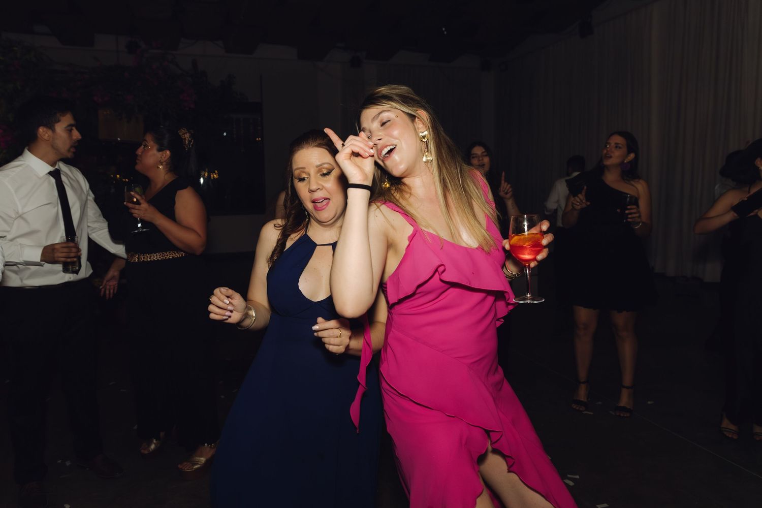 Two women in pink and navy dresses laugh and dance together at a wedding reception.