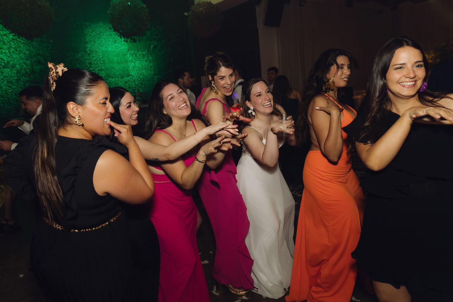 Bride in white gown dances joyfully with bridesmaids in colorful dresses at wedding reception.
