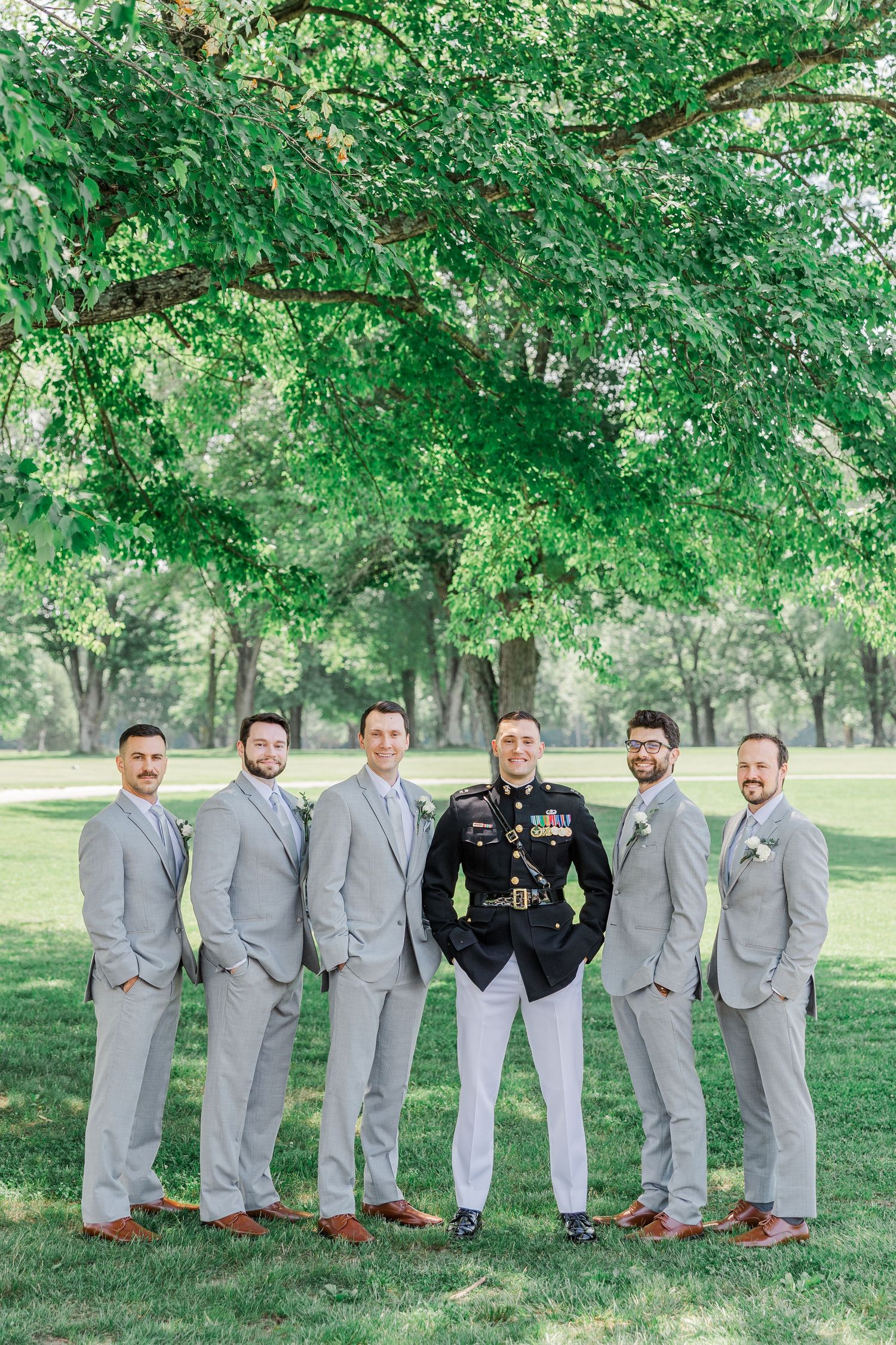 Groom in military dress uniform stands with five groomsmen in gray suits outdoors under lush green trees.