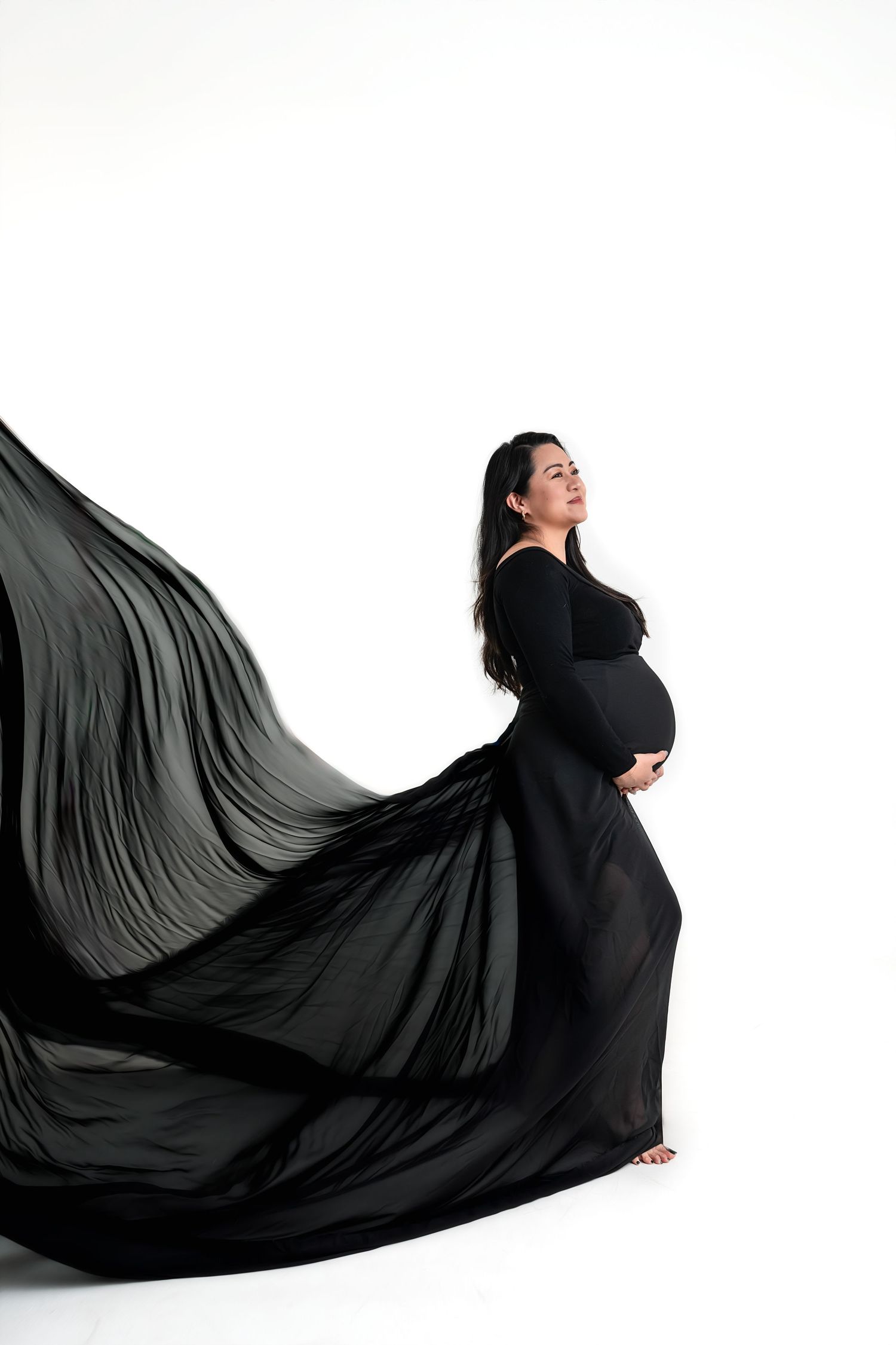 Pregnant woman in flowing black gown cradling baby bump, fabric billowing gracefully against white background.