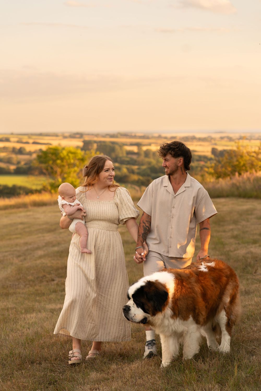 A young family with a newborn baby and Saint Bernard dog enjoying a golden sunset in a scenic countryside field.