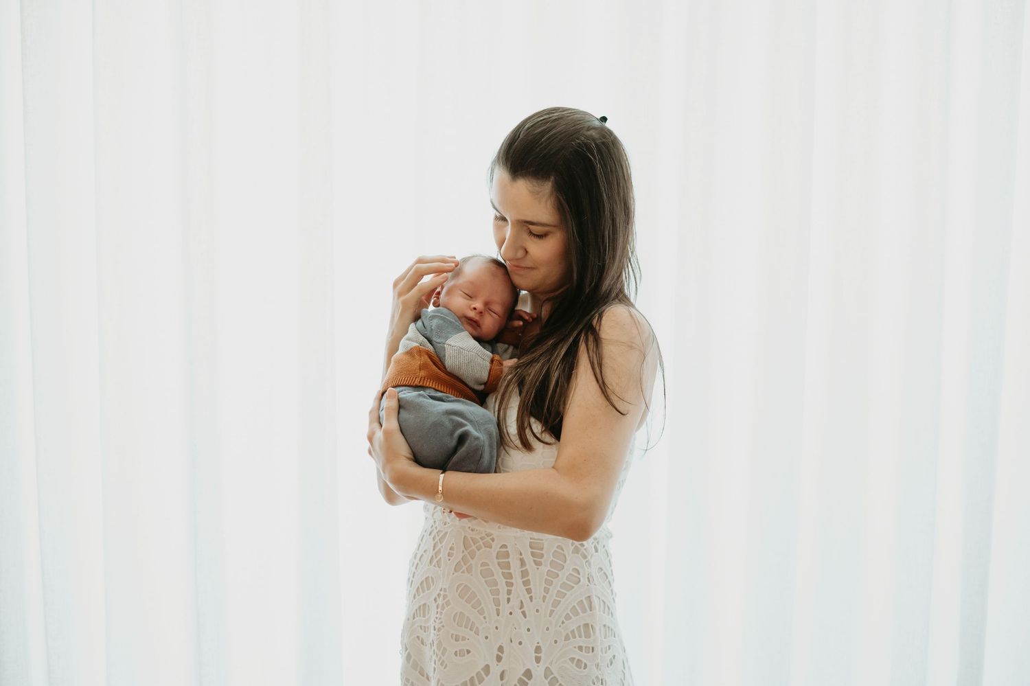 A mother in a white lace dress tenderly holds her newborn baby against a soft white curtain backdrop.