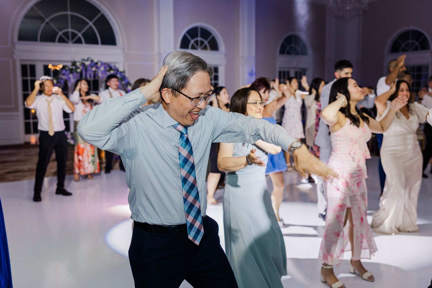 Guests dancing joyfully at a wedding reception in an elegant ballroom with floral decorations.