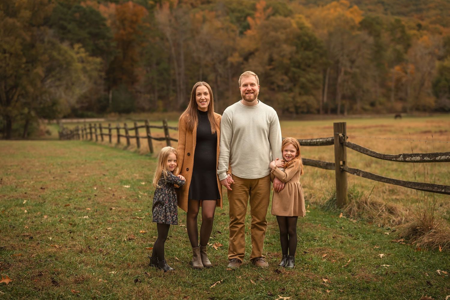 Family of four posing outdoors in autumn field with wooden fence and colorful fall foliage backdrop.