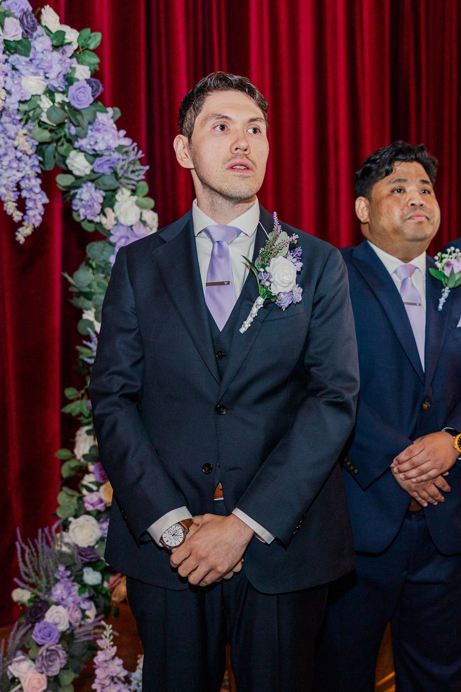 Groom in navy suit with lavender boutonniere stands at wedding altar beside groomsman, with purple floral arrangements nearby.