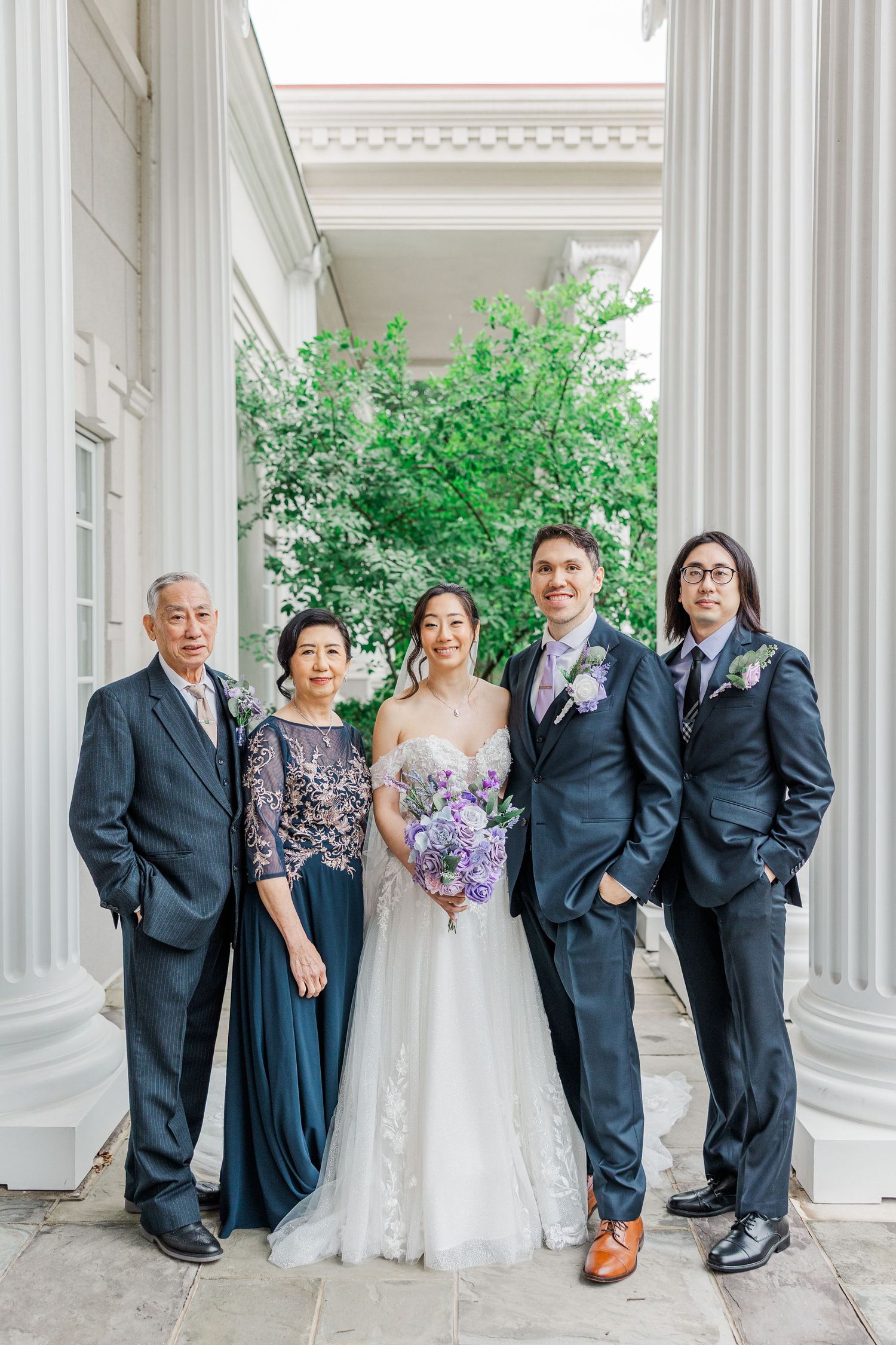 Bride and groom pose with family between white columns, bride holding purple bouquet in elegant lace wedding gown.