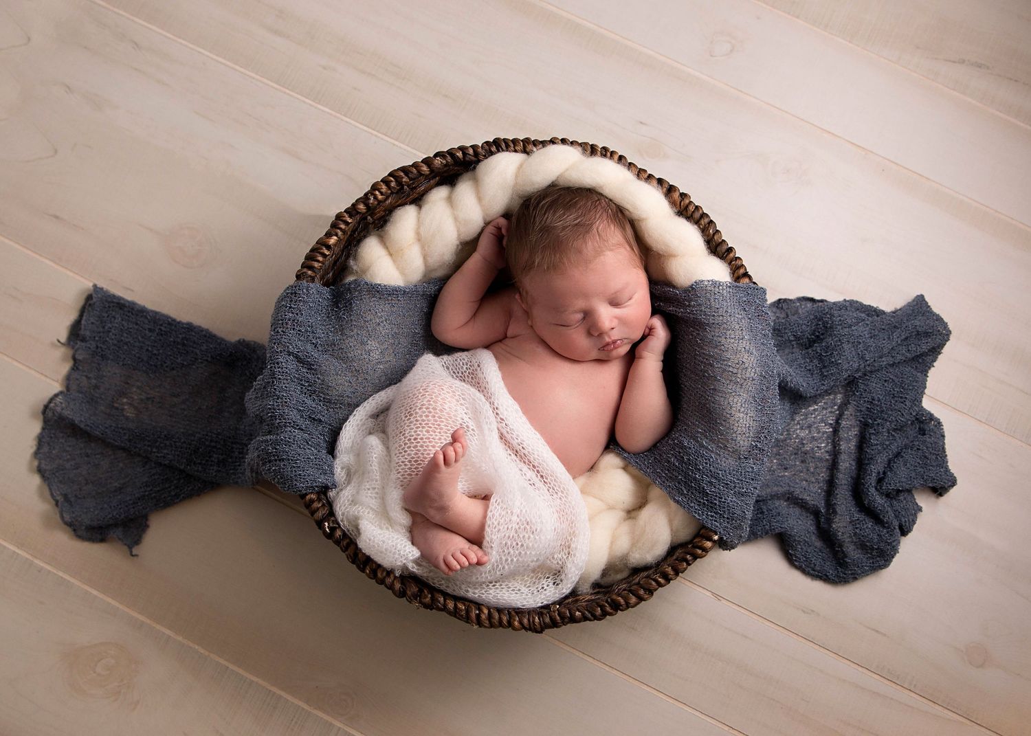 Newborn baby sleeping peacefully in a wicker basket lined with white fur and blue knit wrap on a light wood floor.
