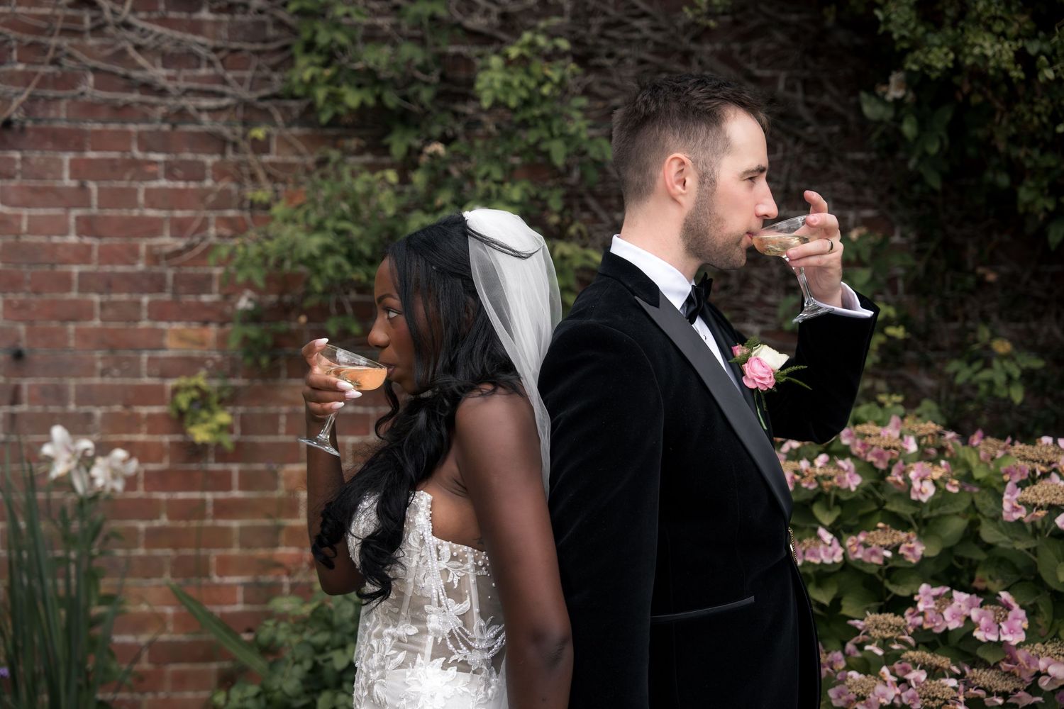 Interracial wedding couple standing back-to-back sipping champagne in garden with brick wall backdrop.
