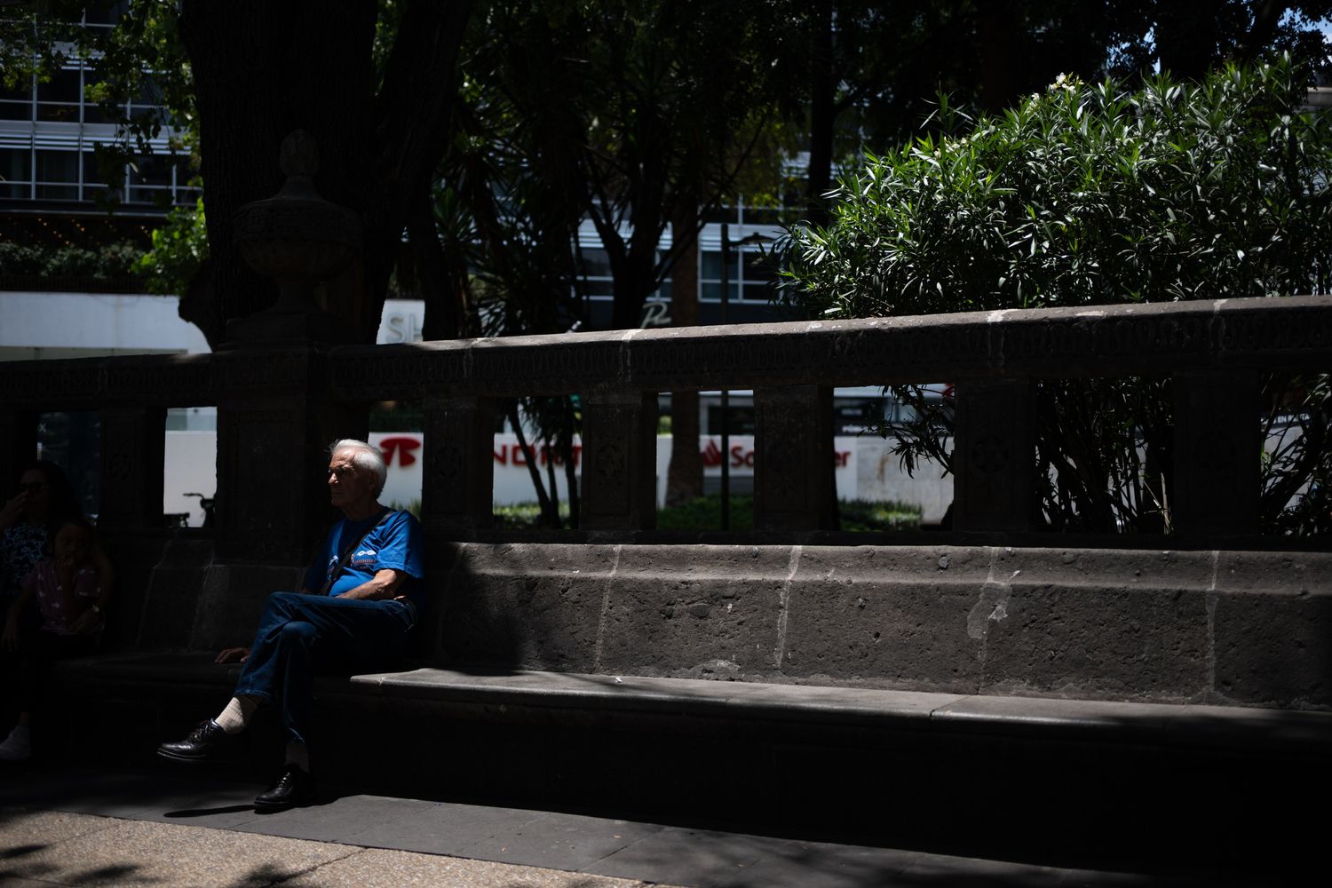 Elderly person sitting alone on a stone bench under a shady tree in an urban park on a sunny day.