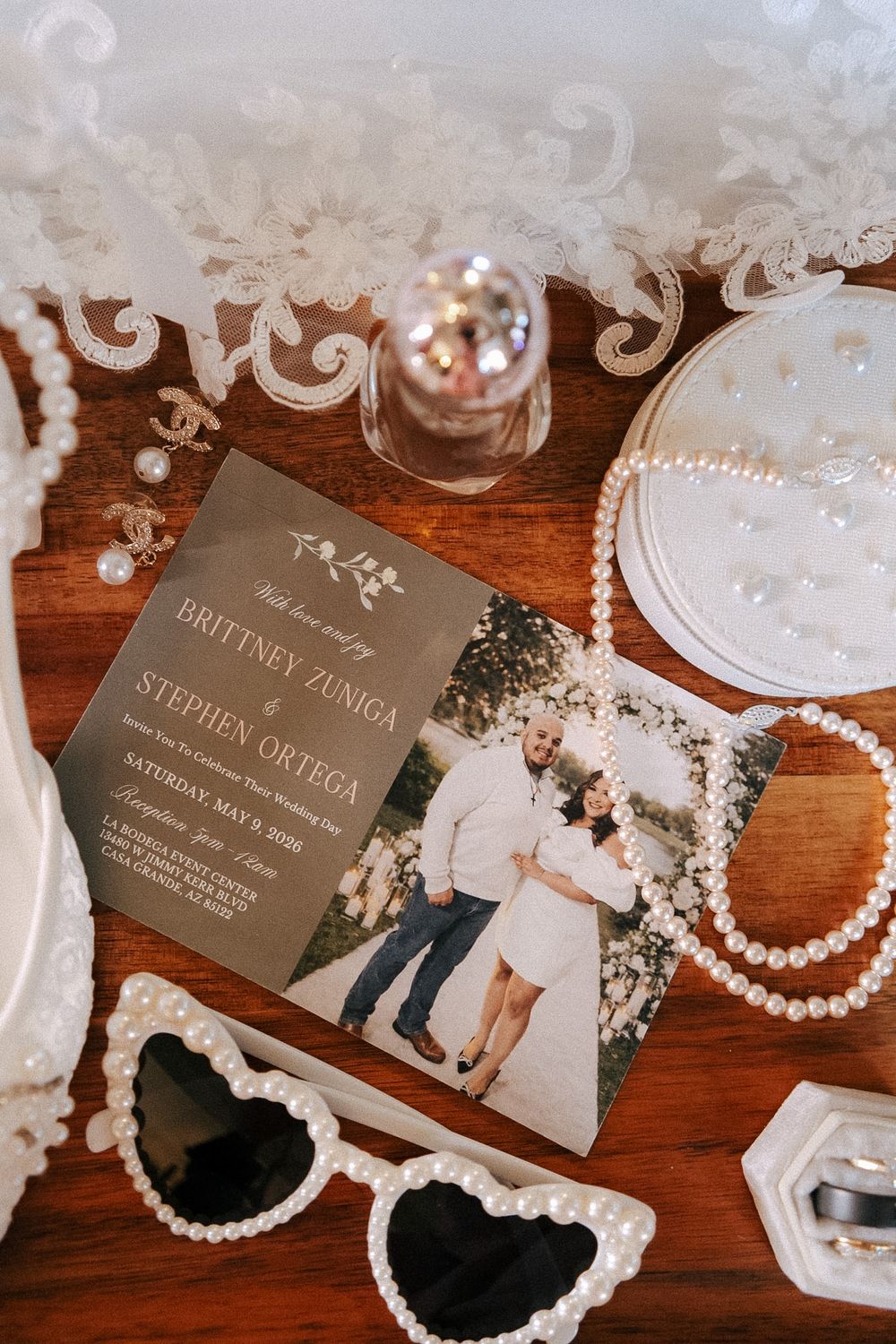 Wedding flat lay featuring invitation for Brittney Zuniga and Stephen Ortega, pearl jewelry, heart sunglasses, and lace details.