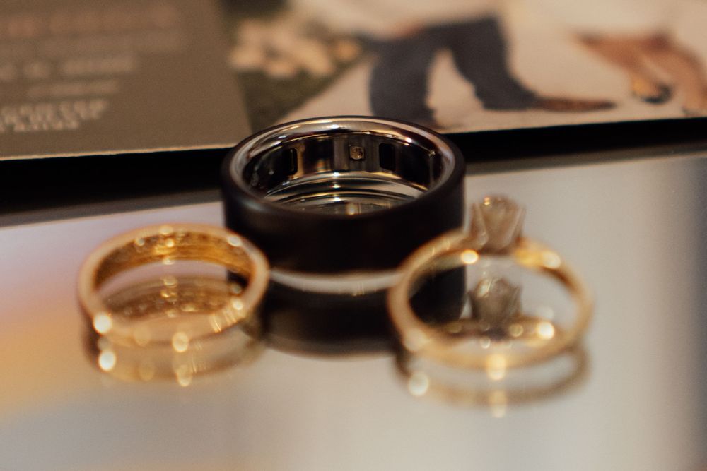 Three wedding rings displayed on a reflective surface, including a black band and two gold rings with diamonds.