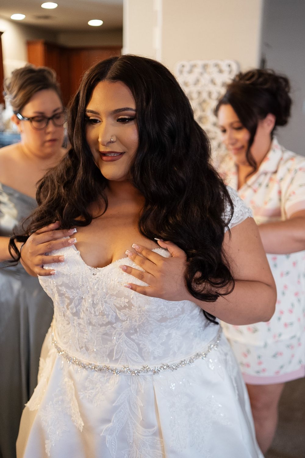 Bride in white lace wedding gown being helped by bridesmaids while adjusting her strapless dress before ceremony.