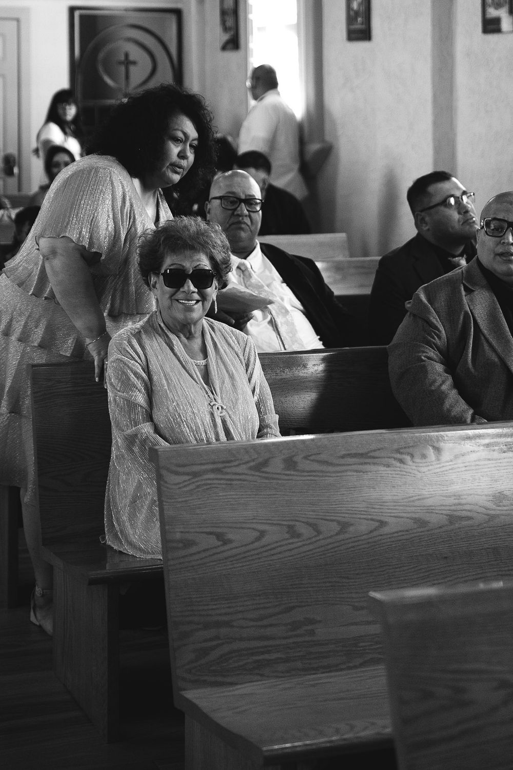 Black and white photo of congregation members seated in church pews, elderly woman in sunglasses smiling during a service.