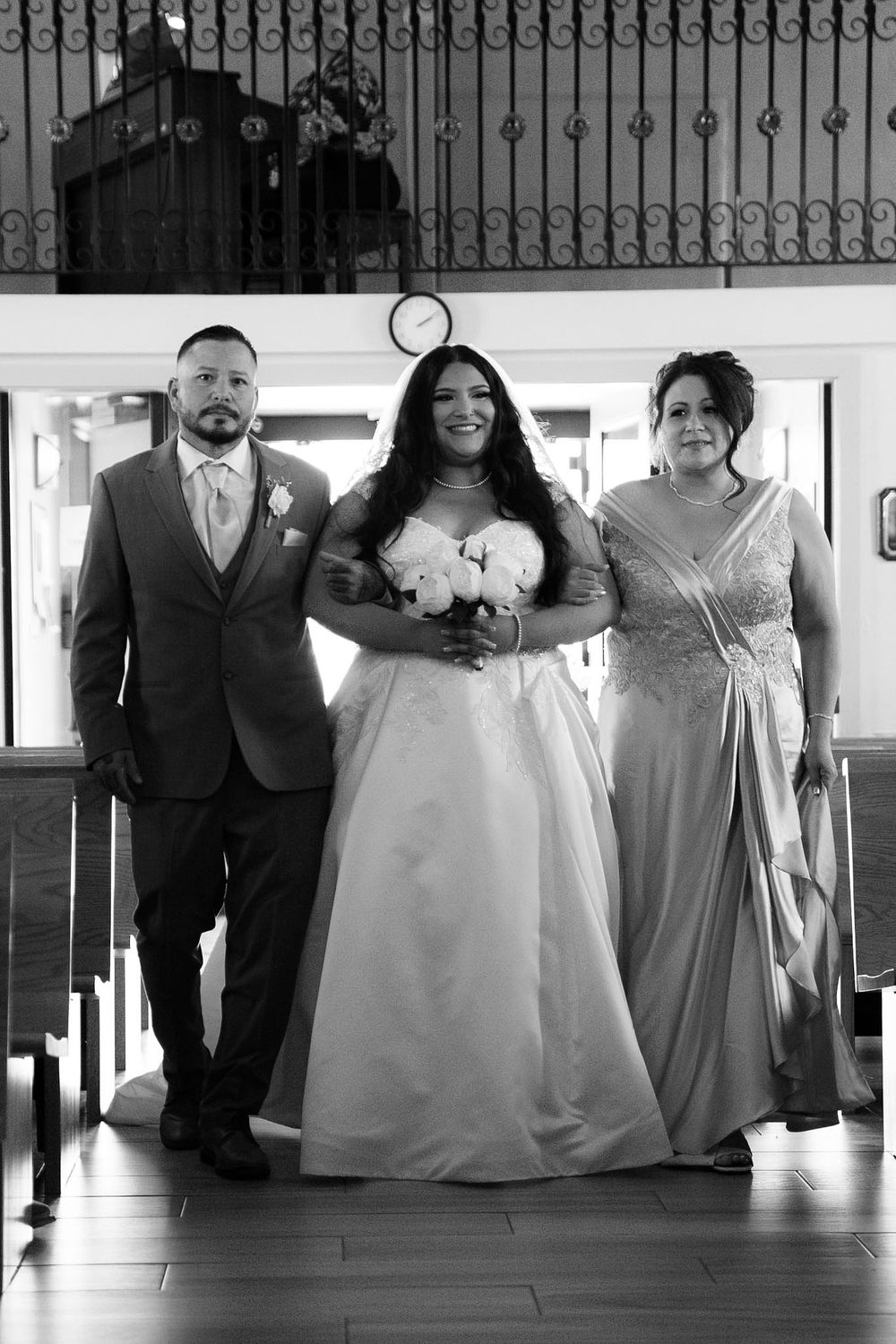 Black and white photo of a bride in a white gown holding bouquet, escorted down the aisle by two family members.