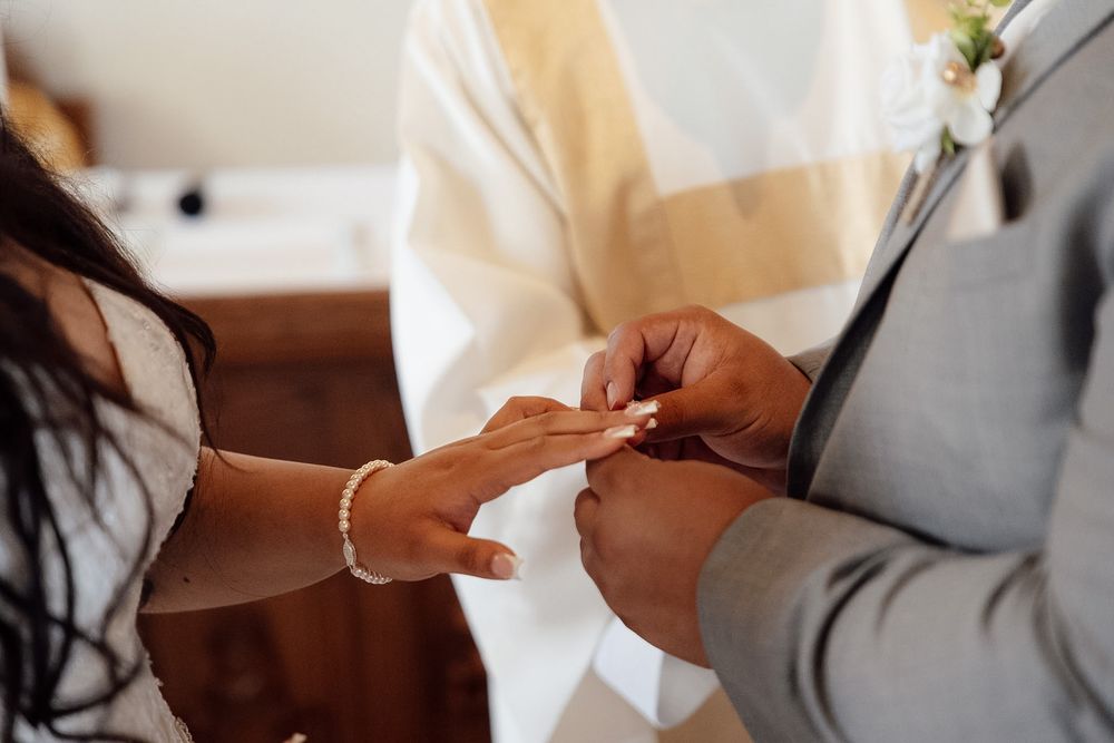 Bride placing a wedding ring on groom's finger during a ceremony, officiant in white robe standing behind.