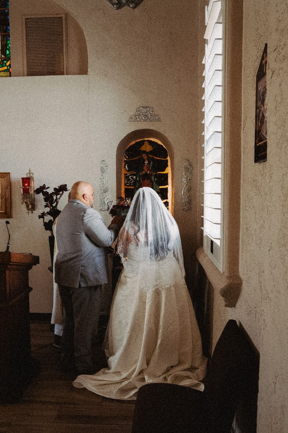 Bride in white gown and veil kneels with groom in gray suit at church altar during wedding ceremony.