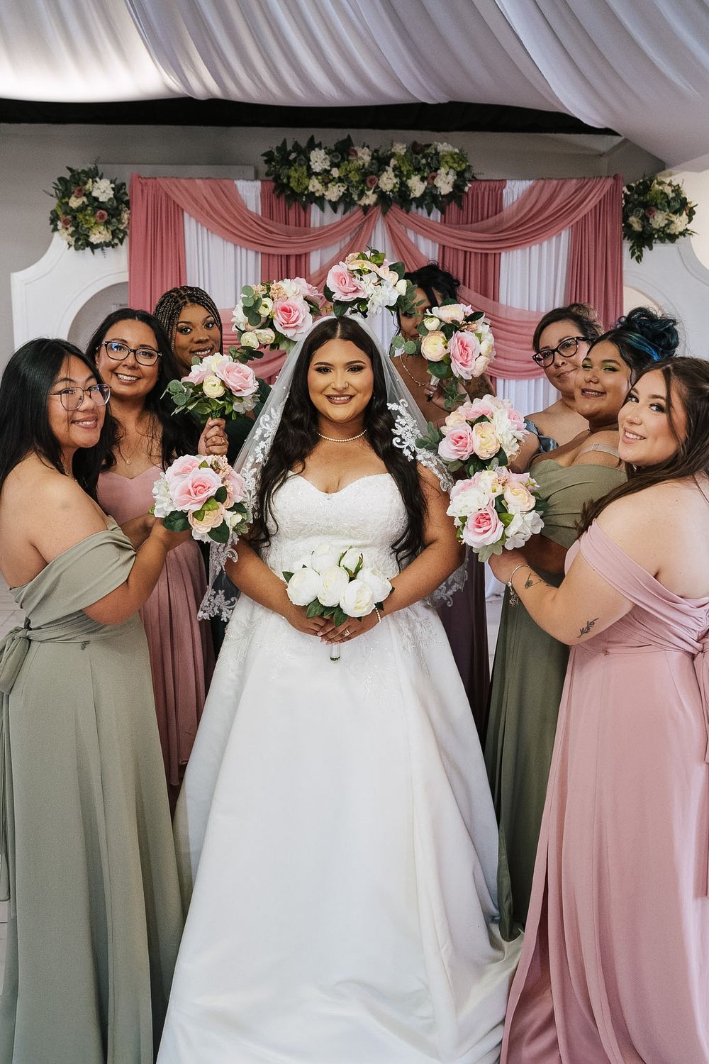 Bride in white gown poses with bridesmaids in sage and pink dresses, holding floral bouquets before a pink draped backdrop.