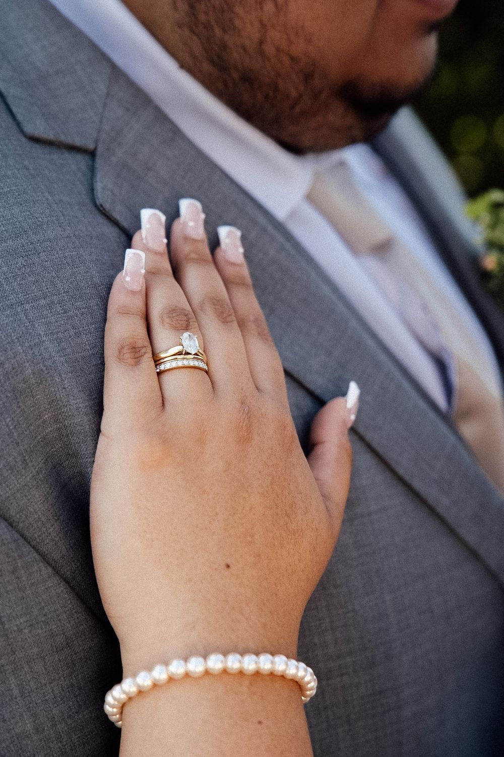 Close-up of bride's hand with pearl bracelet, gold wedding rings, and French manicure resting on groom's gray suit.