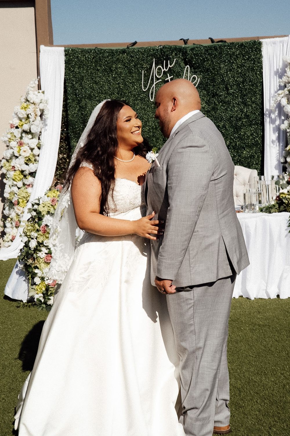 Bride in white gown and groom in gray suit share a joyful moment at outdoor wedding ceremony with floral backdrop.