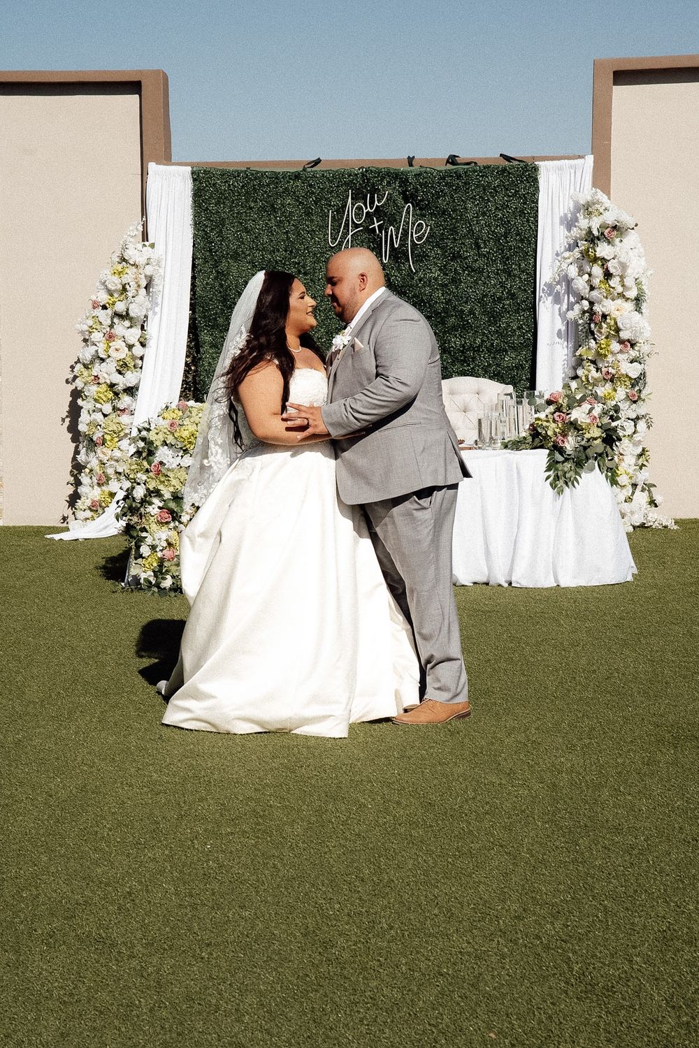 Bride and groom share a romantic moment at outdoor wedding ceremony with floral arch and green hedge backdrop.