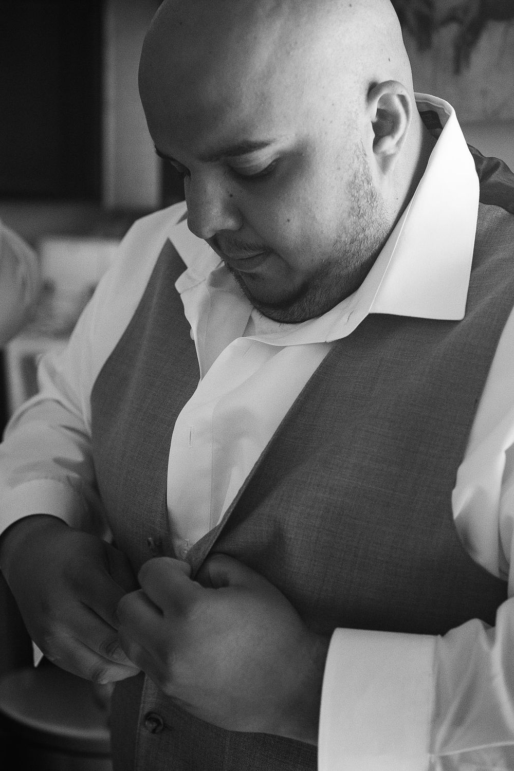 Black and white photo of a bald man in a suit vest buttoning his shirt before a formal event.