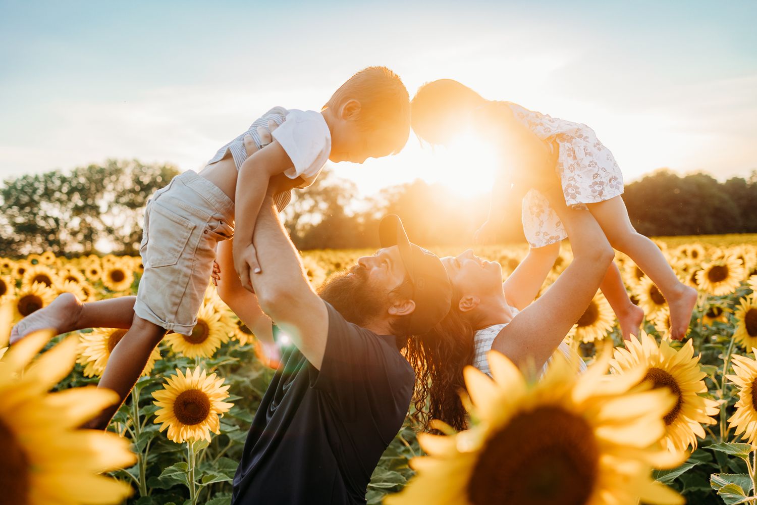 Image taken in the sunflowers, mom and dad holding the kids up while they are laughing and smiling. The sun shines in the background.