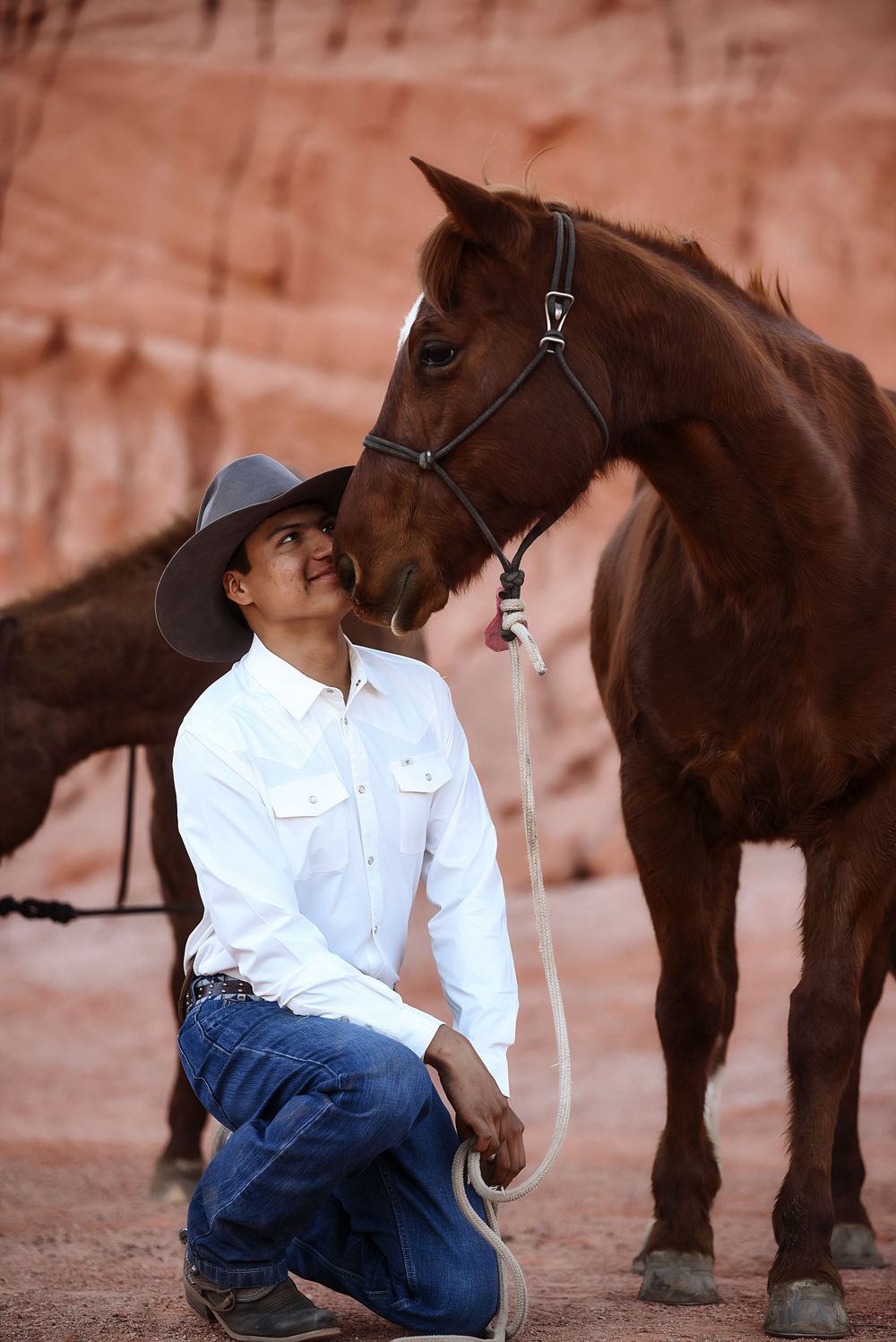 Cowboy in white shirt and hat kneels beside a chestnut horse in a red rock canyon setting.