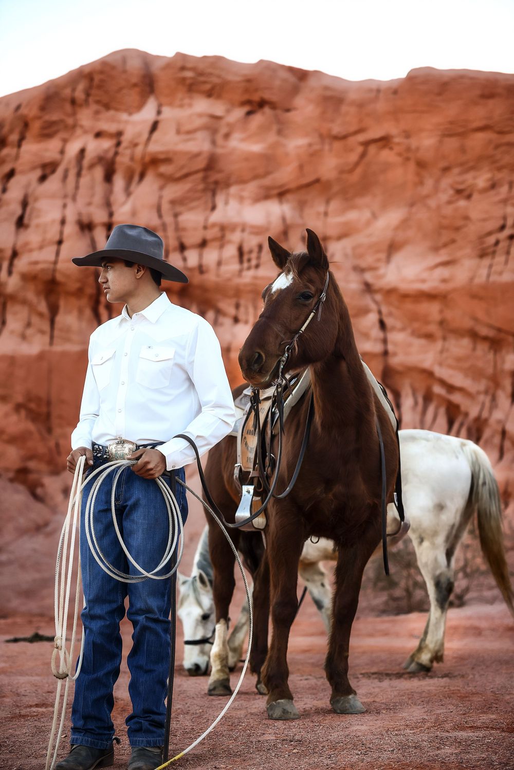 Cowboy in white shirt and black hat holds lasso beside brown horse against red canyon walls.