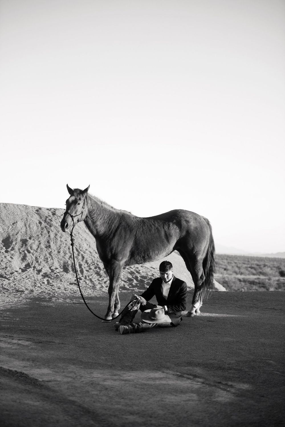 Black and white photo of a man sitting beneath a horse on a desert road, holding its lead rope.