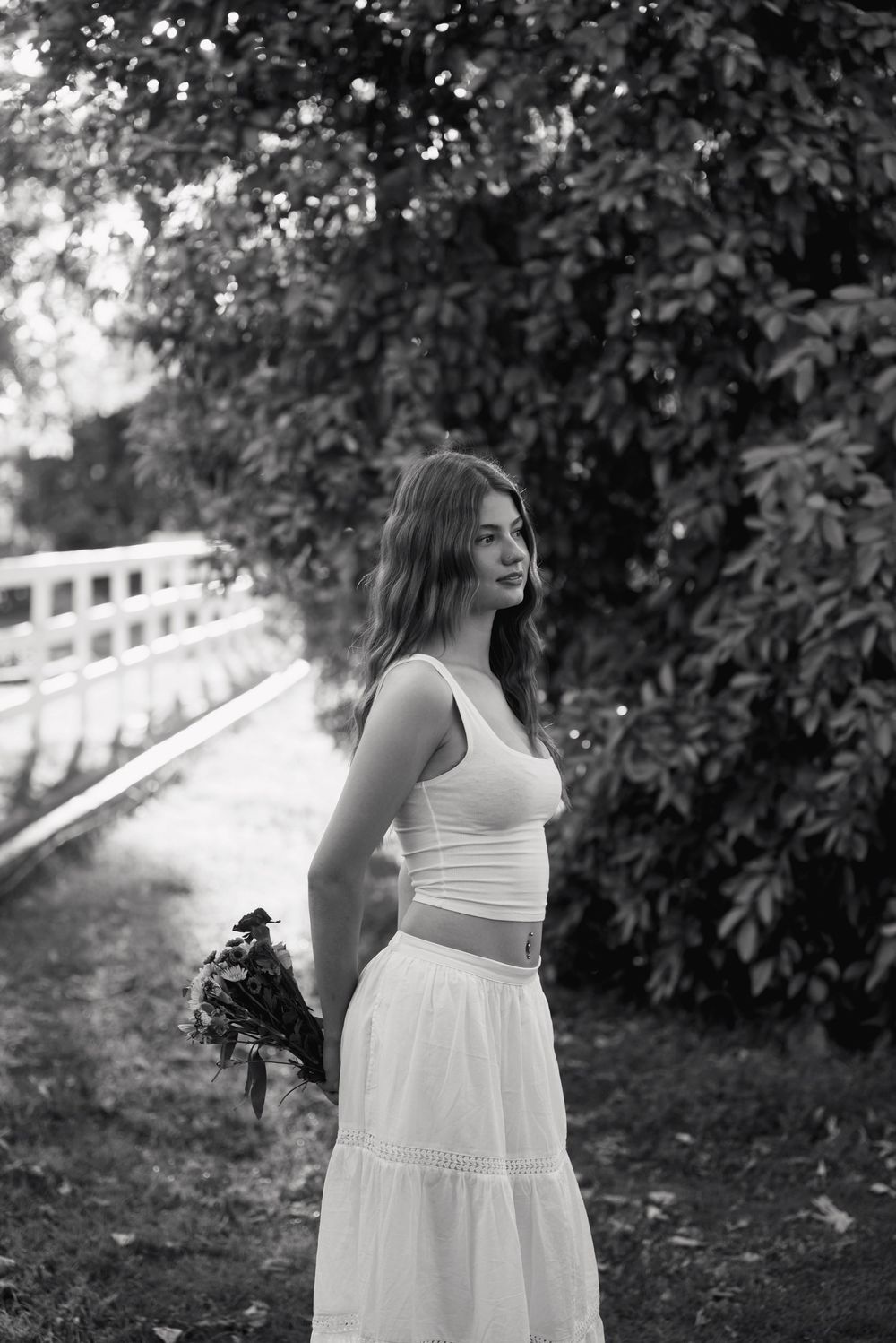 Black and white photo of a young woman in white crop top and maxi skirt holding flowers outdoors near trees.
