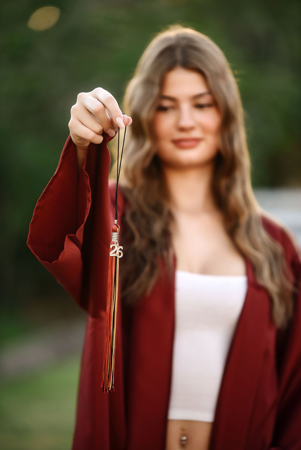 Young woman in maroon graduation gown holds tassel toward camera in outdoor portrait photo.