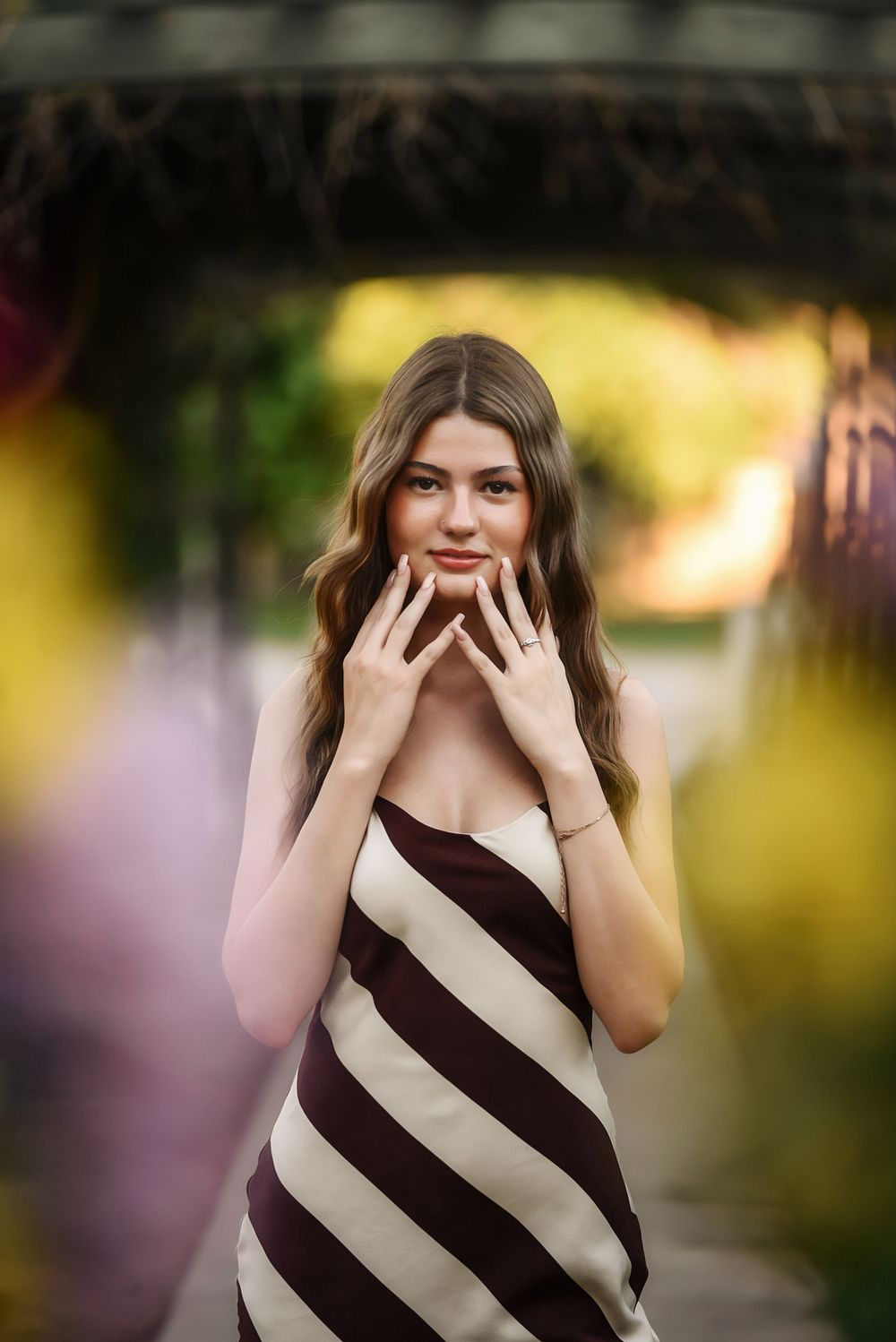 Young woman in striped brown and white dress posing outdoors among colorful bokeh flowers.