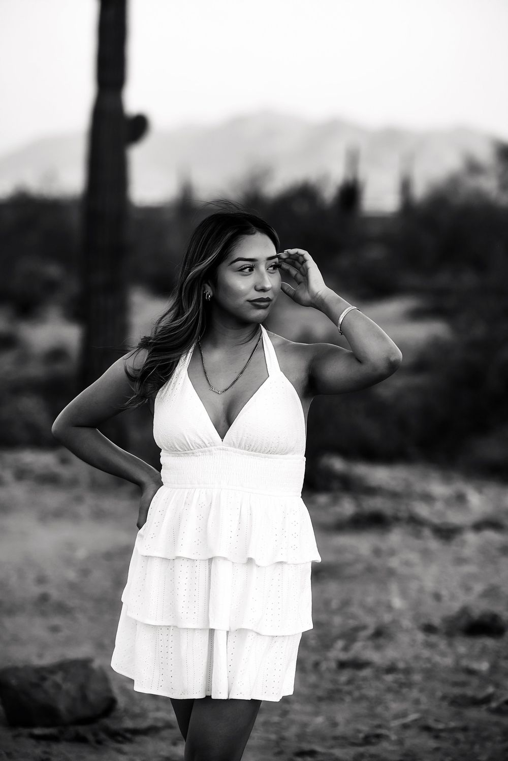 Black and white photo of a young woman in a white ruffled halter dress posing outdoors with cacti and mountains behind her.
