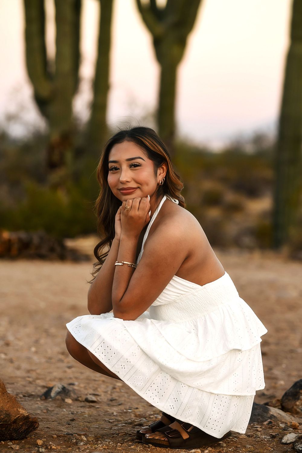 Young woman in white halter dress crouching in desert landscape with saguaro cacti at sunset.