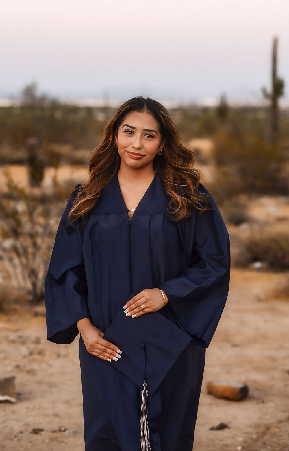 Young woman in navy graduation cap and gown posing outdoors in desert landscape with cacti at dusk.