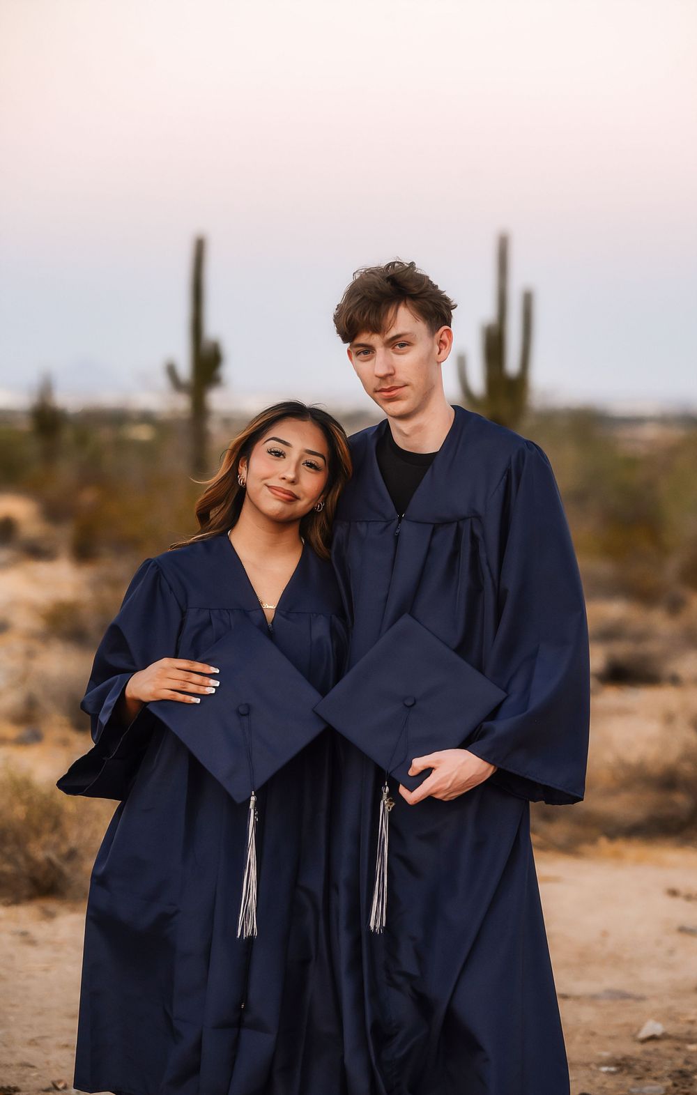 Two graduates in navy blue caps and gowns pose together in a desert landscape with saguaro cacti at dusk.