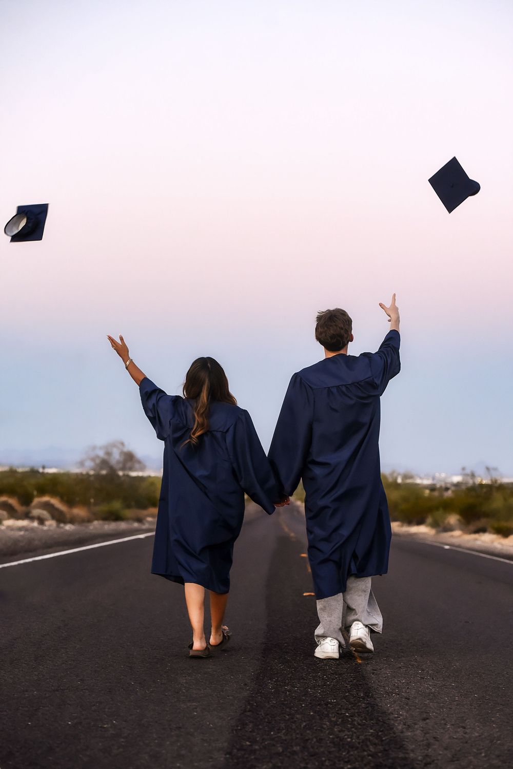 Two graduates in navy blue gowns toss caps in the air while holding hands on an open road at dusk.