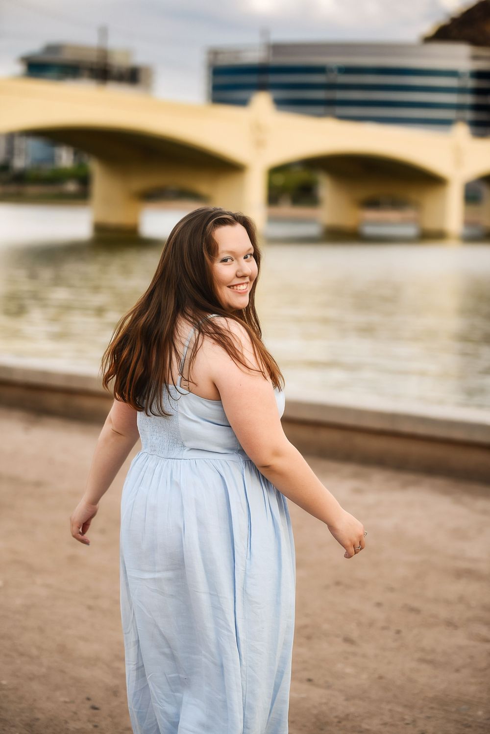 Young woman in light blue sundress smiling by a river with a yellow bridge and city buildings in the background.