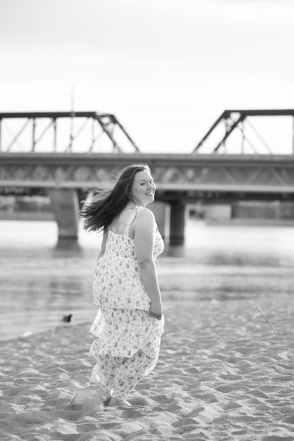 Black and white photo of a smiling woman in a floral dress on a sandy beach with a bridge in the background.