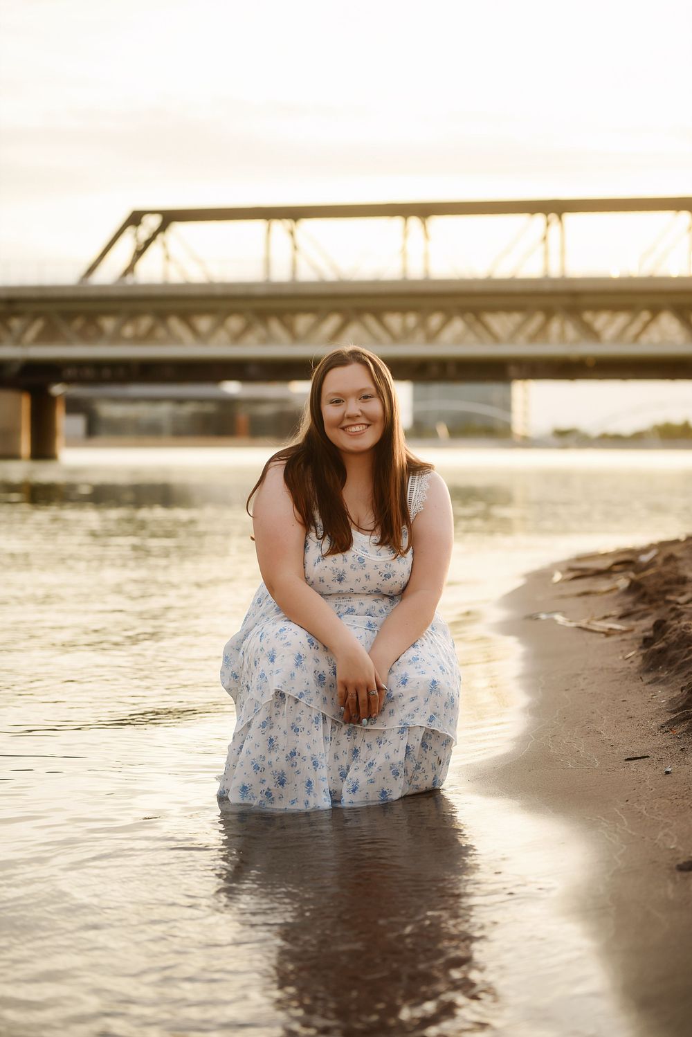 Young woman in floral dress sitting by river shore at golden hour with bridge in background.