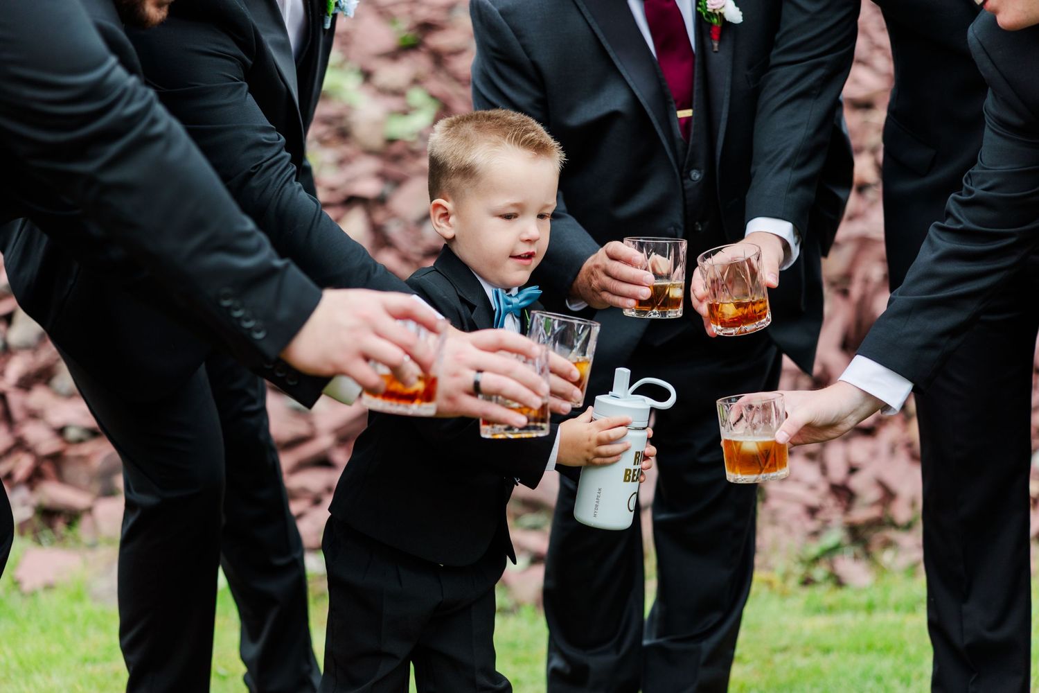 Young ring bearer in black suit toasting drinks with groomsmen at an outdoor wedding celebration.