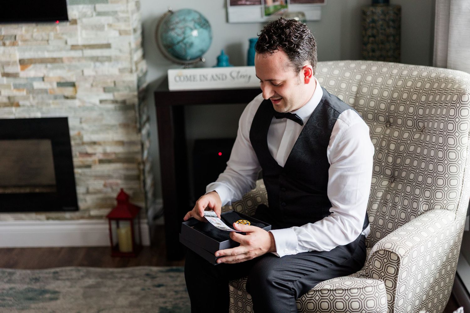 Groom in black vest and bow tie sits in armchair, examining wedding gift box before ceremony.