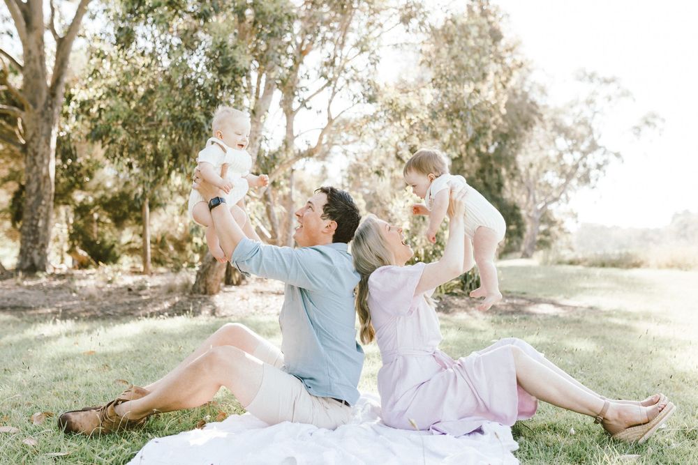 A happy family sits on a blanket in a sunny park, parents lifting two babies in the air outdoors.