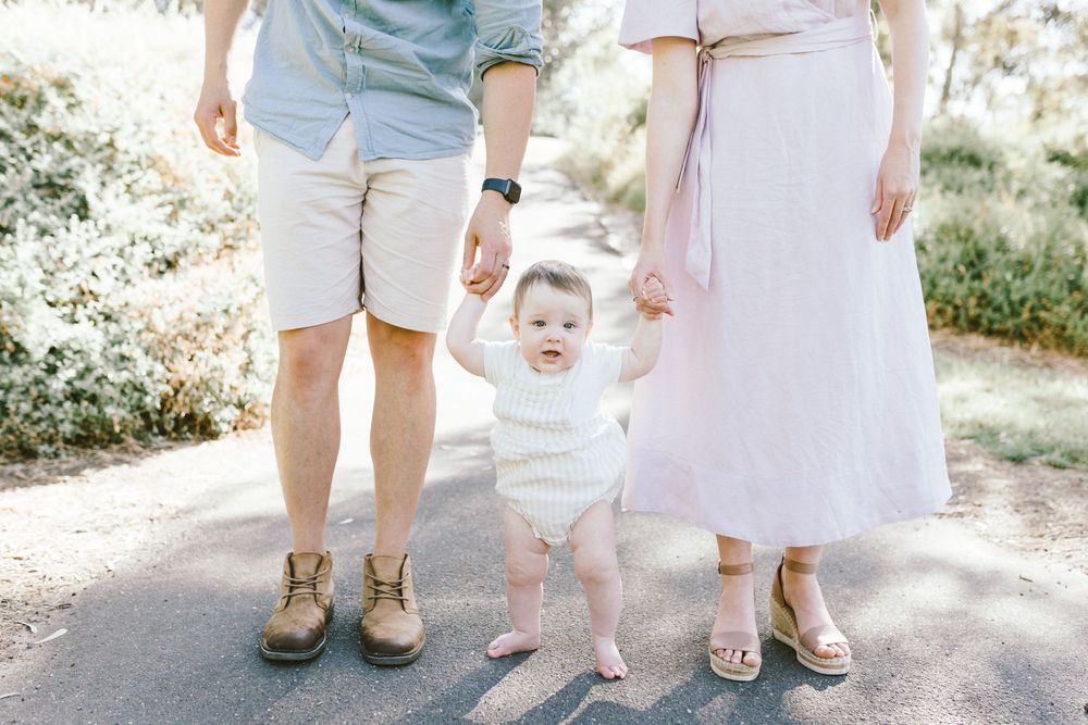 Parents holding hands with their baby taking first steps on a sunny garden path, family outdoor portrait.